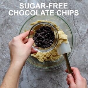 Top view photo of Keto Cookie Dough in a glass bowl, with a hand adding chocolate chips to the dough.