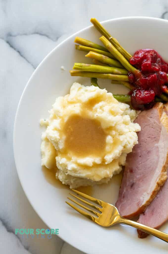 a white plate with gravy on mashed cauliflower, slices of ham. cranberry jelly and asparagus.