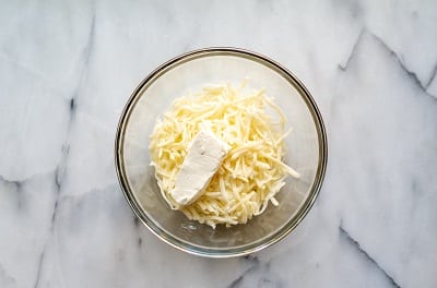 Top view photo of a glass bowl with shredded mozzarella cheese and cream cheese in the bowl, ready to be mixed.