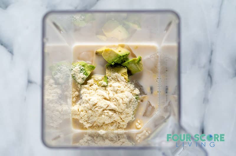 ingredients for a smoothie including avocado, protein powder, ice, and milk in a blender