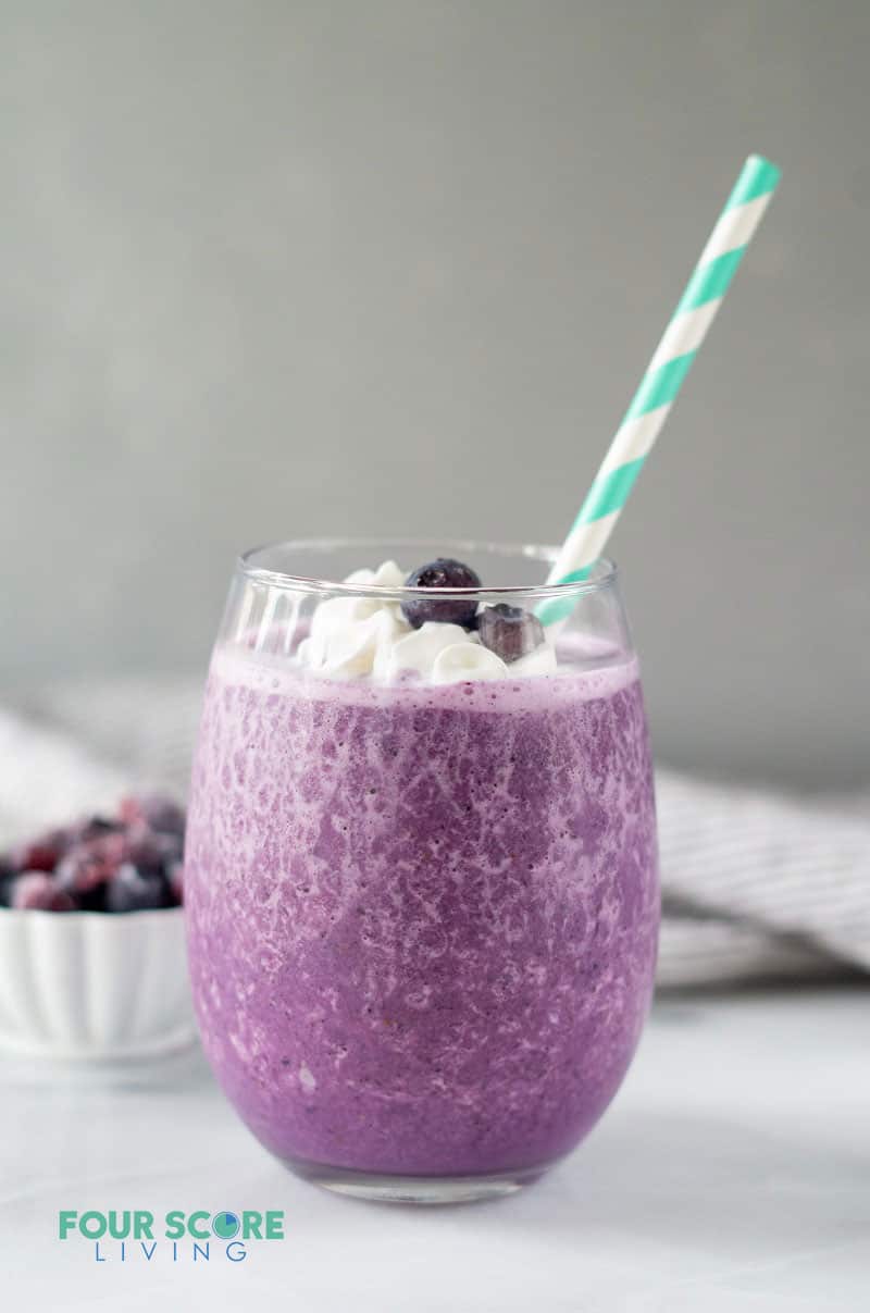 a purple colored keto berry smoothie topped with whipped cream and blueberries in a clear glass with a blue and white straw
