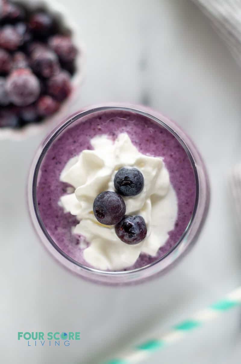 a glass filled with a purple colored frozen keto berry smoothie and topped with white cream and three blueberries