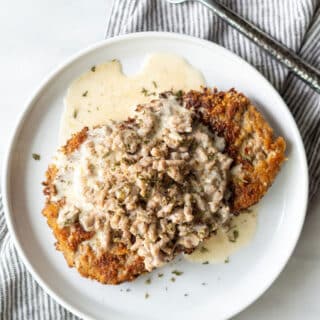 close up of chicken fried steak with gravy on a round white plate next to a fork and a striped napkin