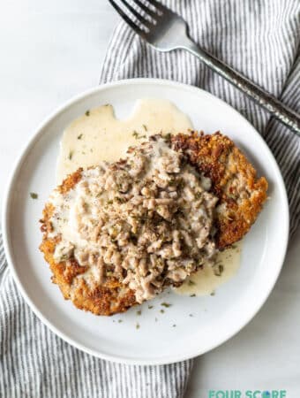 close up of chicken fried steak with gravy on a round white plate next to a fork and a striped napkin