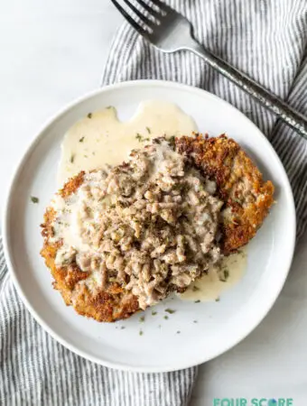 close up of chicken fried steak with gravy on a round white plate next to a fork and a striped napkin