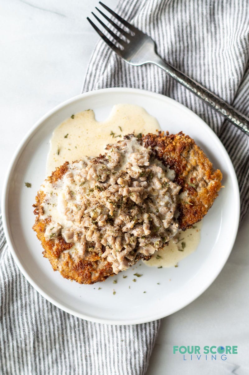 chicken fried steak with gravy on a round white plate next to a fork and a striped napkin