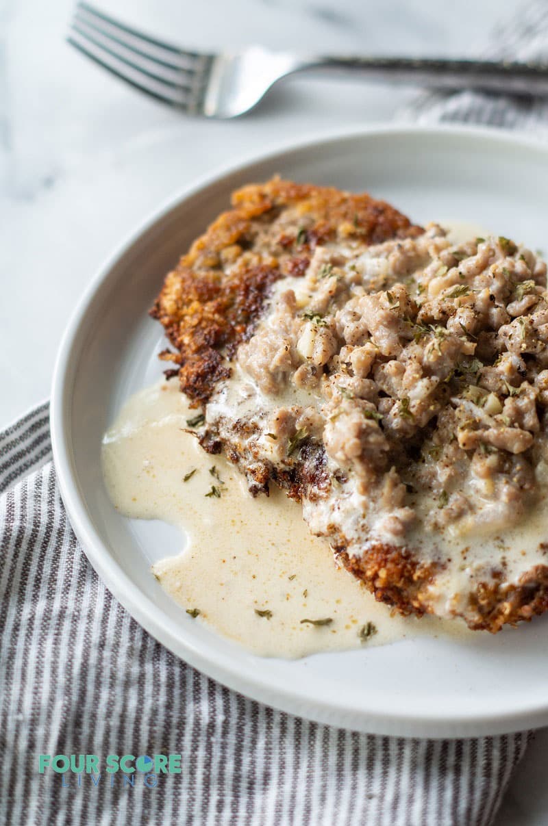 a side view of chicken fried steak with gravy on a round white plate next to a fork and a striped napkin