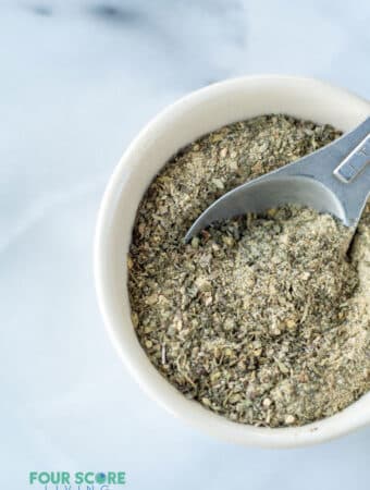 a measuring spoon scooping into a round white bowl of a greenish seasoning mix