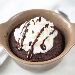 a chocolate mug cake with whipped cream and a drizzle of fudge in a brown ramekin