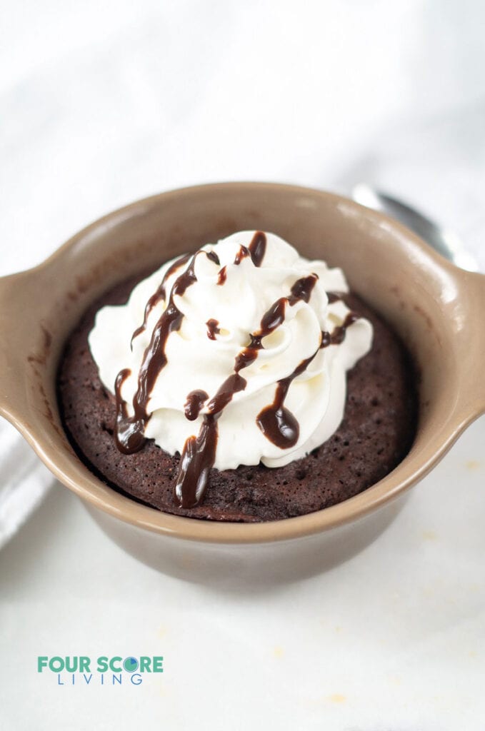 a chocolate mug cake with whipped cream and a drizzle of fudge in a brown ramekin