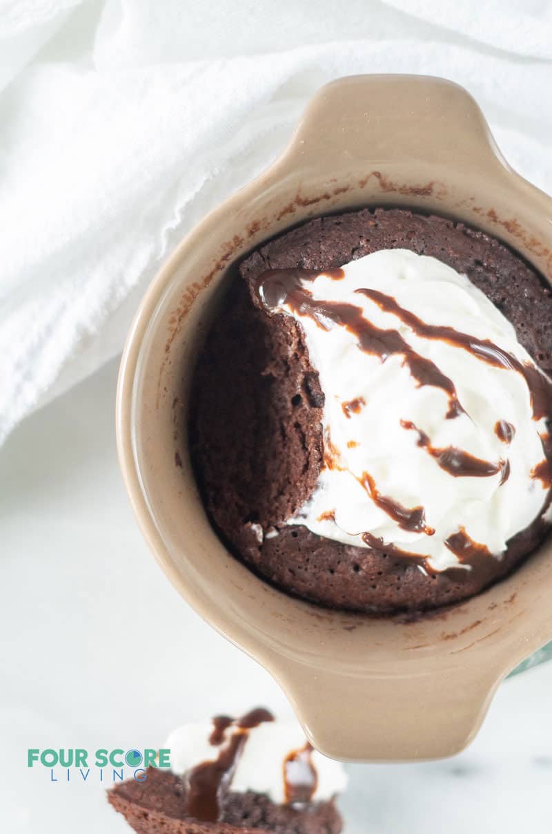 a chocolate cake topped with whipped cream and chocolate sauce in a brown ramekin with a bite taken from the side of the cake