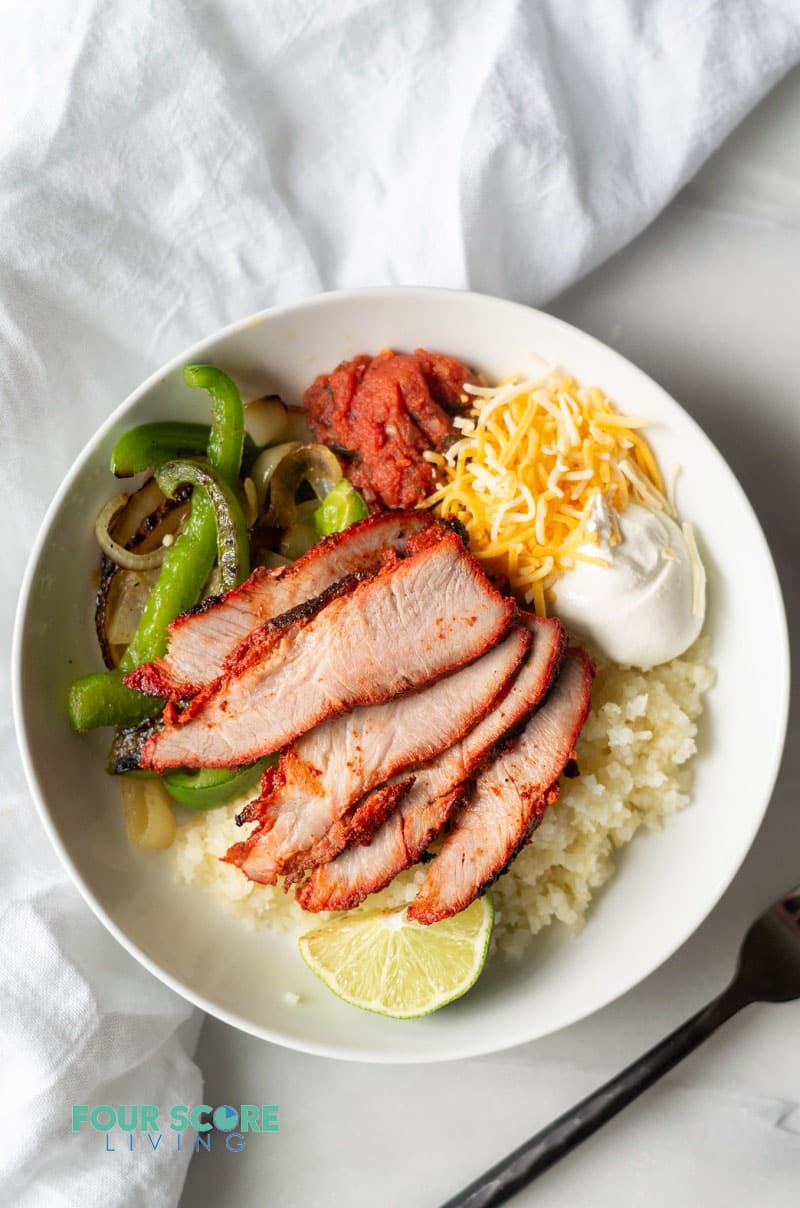 a white round bowl filled with slices of spiced pork asada, green peppers, cheese, sour cream, salsa, rice, and a lime wedge