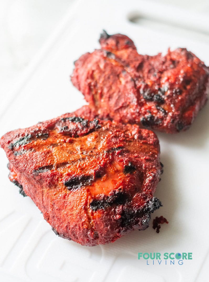 filets of cooked spiced pork carne asada with red-colored seasoning and black grill marks on a white cutting board