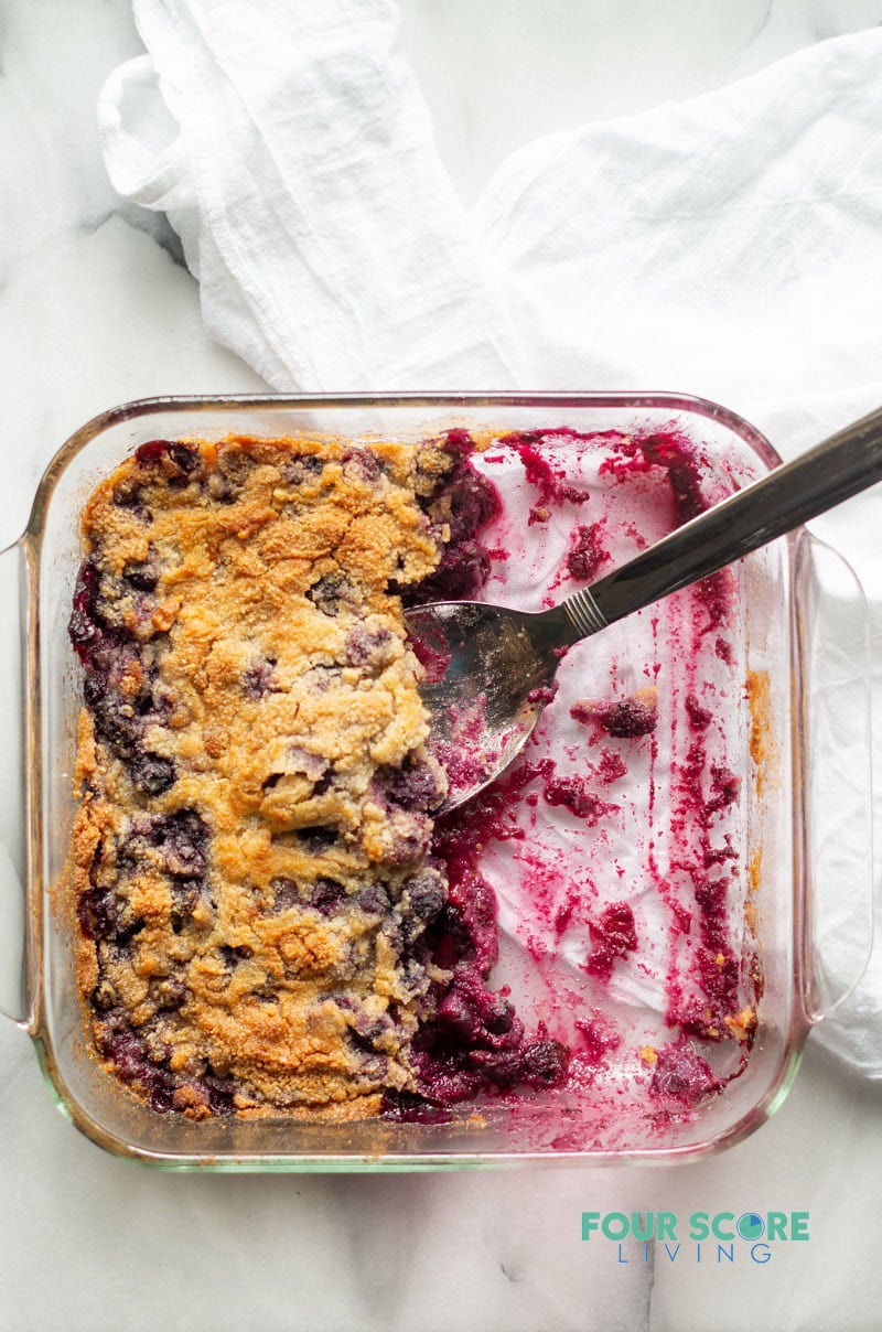 half of a blueberry dump cake remaining in a square glass baking dish with a silver serving spoon