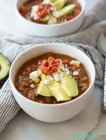 a side view of keto taco soup in a white bowl, topped with avocado and salsa