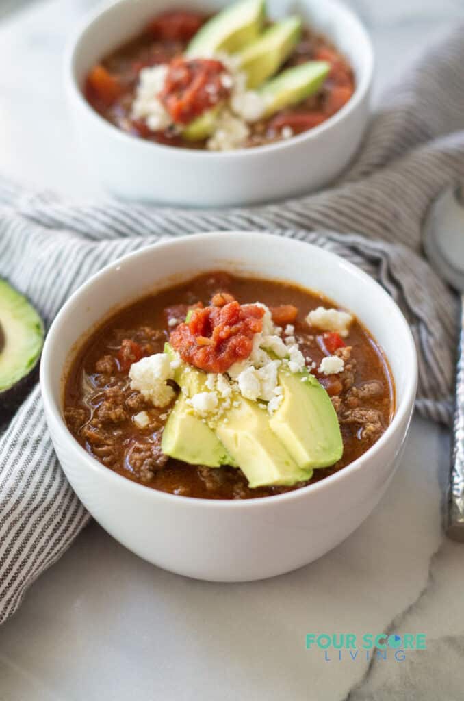 a side view of keto taco soup in a white bowl, topped with avocado and salsa