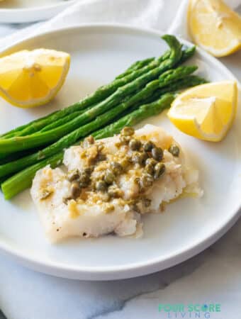 Cod topped with capers and garlic served with lemon wedges and asparagus on a white plate.