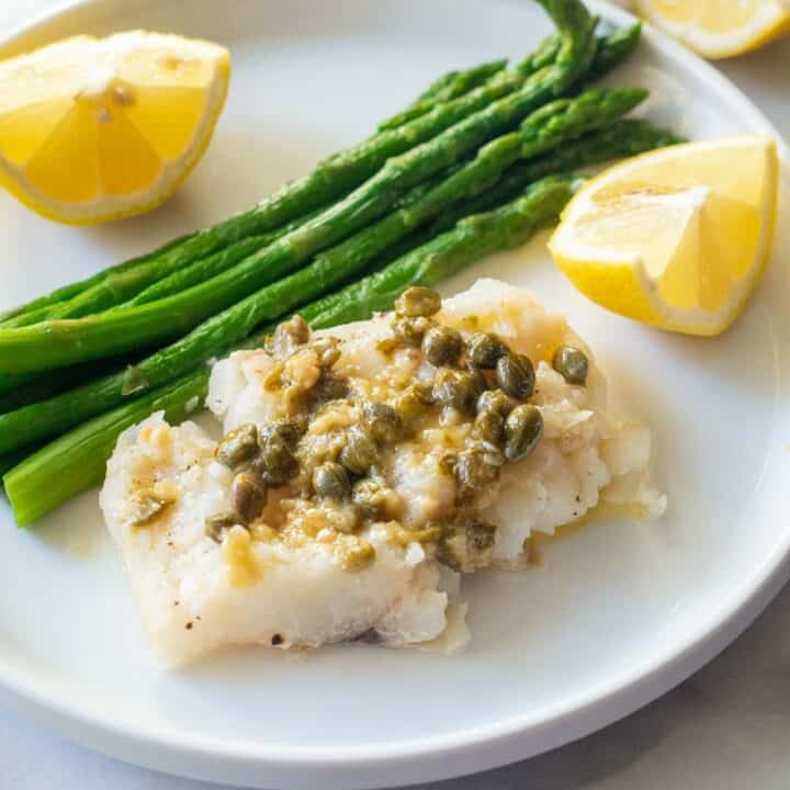 Cod topped with capers and garlic served with lemon wedges and asparagus on a white plate.