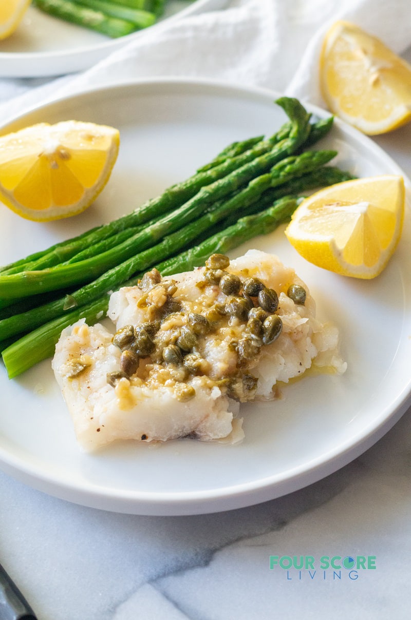 Cod topped with capers and garlic served with lemon wedges and asparagus on a white plate.