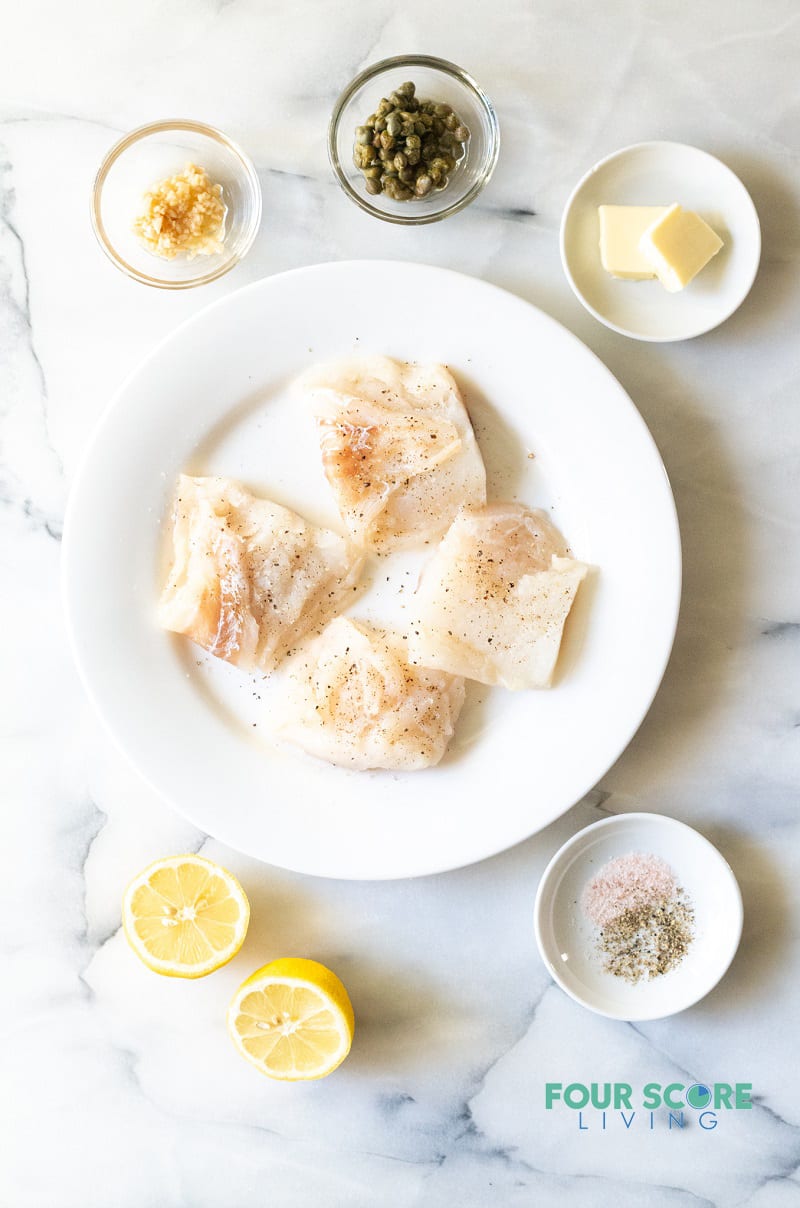 Top down image of ingredients for Keto cod. Fish fillets, lemons, capers, garlic, butter, and seasonings in separate dishes.