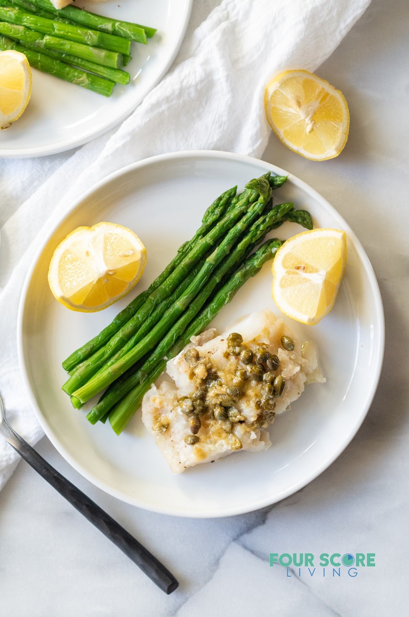 Cod topped with garlic and caper sauce served with lemon wedges and asparagus on a white plate.