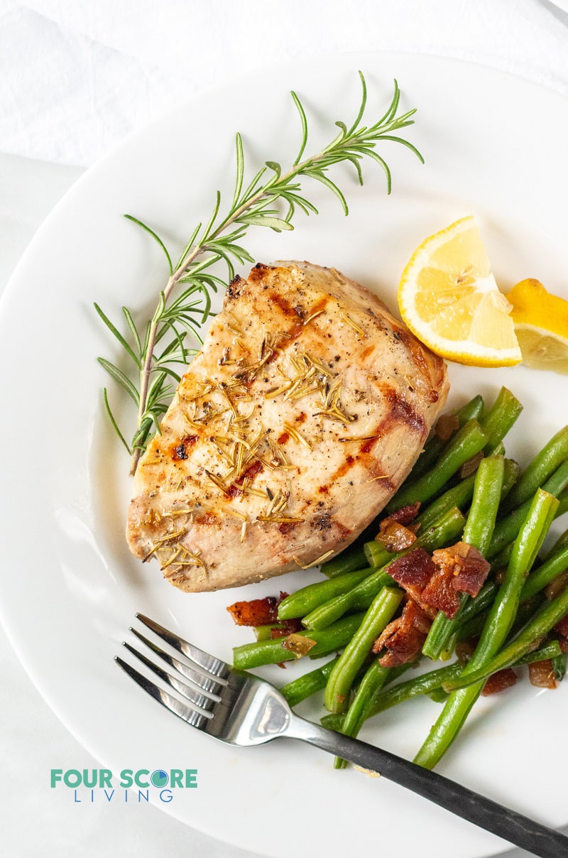 pork chop on a plate served with green beans, lemon wedges, and a rosemary garnish.