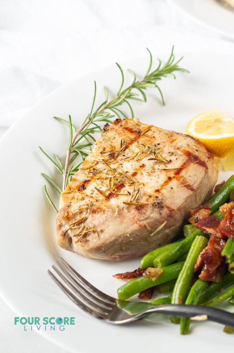 closeup of grilled pork chop on a plate, served with green beans with bacon, garnished with rosemary