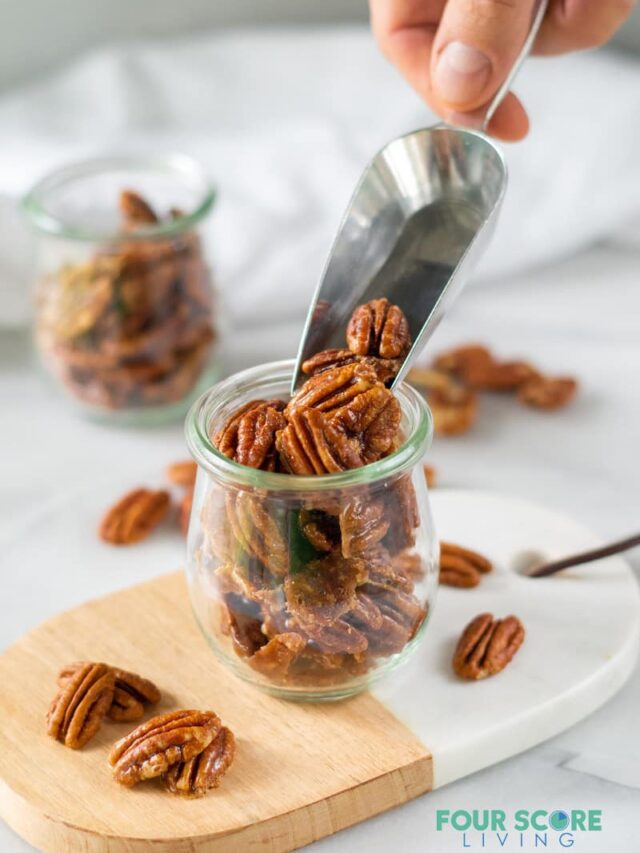 a scoop of keto candied pecans filling a small clear glass jar resting on an oval cutting board made of half wood and half marble, all resting on a white stone surface. 