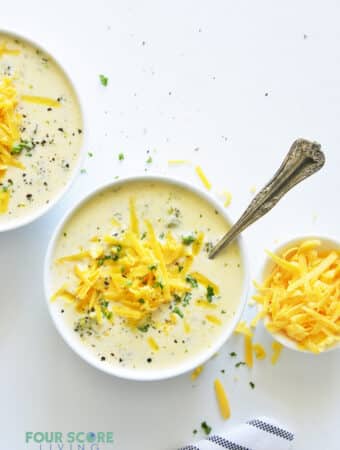 Two bowls of creamy broccoli cheese soup next to a small bowl of shredded cheese