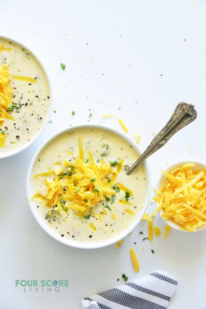 Two bowls of creamy broccoli cheese soup next to a small bowl of shredded cheese