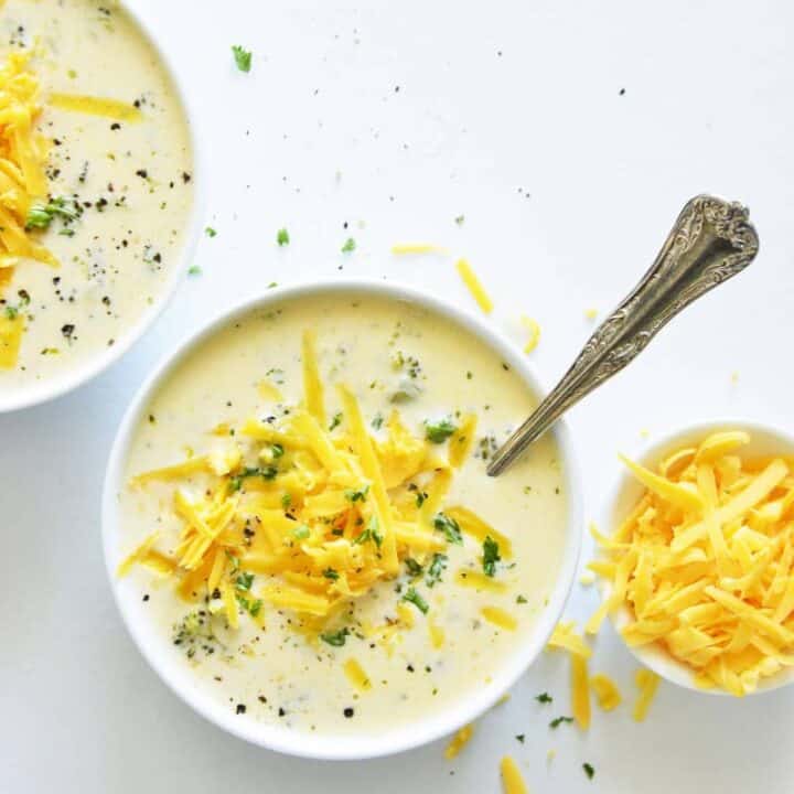Two bowls of creamy broccoli cheese soup next to a small bowl of shredded cheese