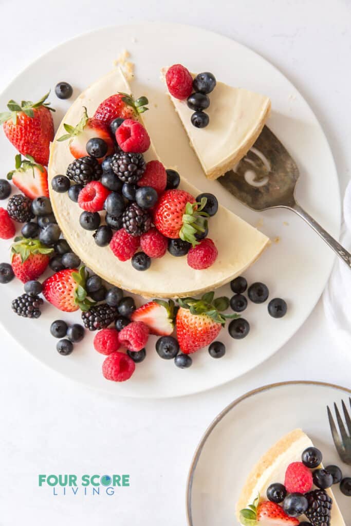 top down view of cheesecake topped with fresh berries. One slice is on a plate, another slice is being served.