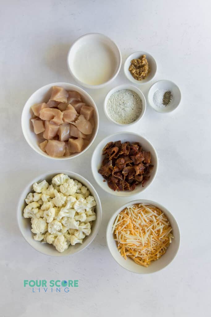 Top down image of small bowls with casserole ingredients, including chicken, cauliflower, cheese, bacon, and seasonings.