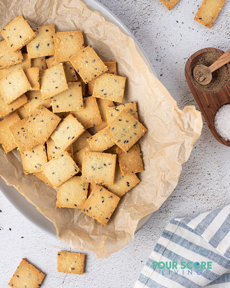 An oval dish lined with brown parchment paper with square crackers inside, and a double wooden spice bowl to the side.