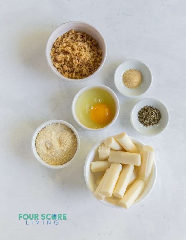 Ingredients for making keto mozzarella sticks, each in separate bowls. Including cheese sticks, pork rinds, egg, and seasoning.