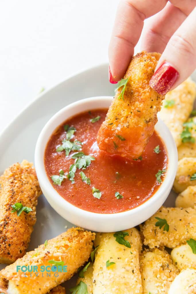 Mozzarella stick being dipped into marinara sauce by a feminine hand.