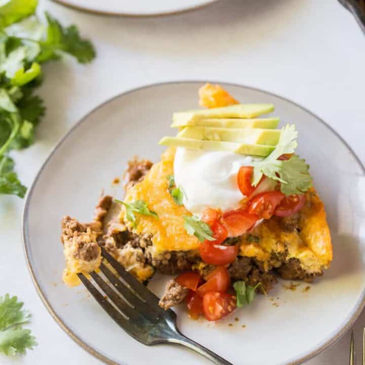 a slice of taco pie on a plate, topped with sour cream, avocoado, tomatoes, and cilantro.