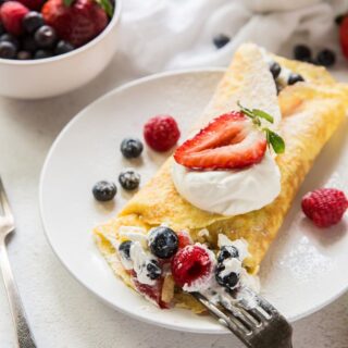 a round white plate with a folded crepe stuffed with cream and mixted berries, being eated with a fork.. Near the plate is a bowl of berries and a bowl of whipped cream