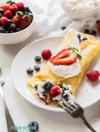 a round white plate with a folded crepe stuffed with cream and mixted berries, being eated with a fork.. Near the plate is a bowl of berries and a bowl of whipped cream