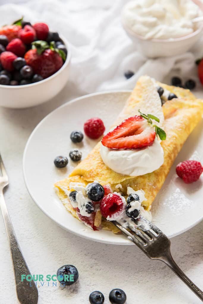a round white plate with a folded crepe stuffed with cream and mixted berries, being eated with a fork.. Near the plate is a bowl of berries and a bowl of whipped cream