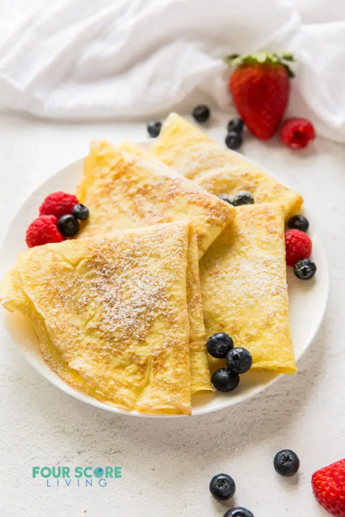 a round white plate topped with crepes folded in triangles topped with raspberries and blueberries