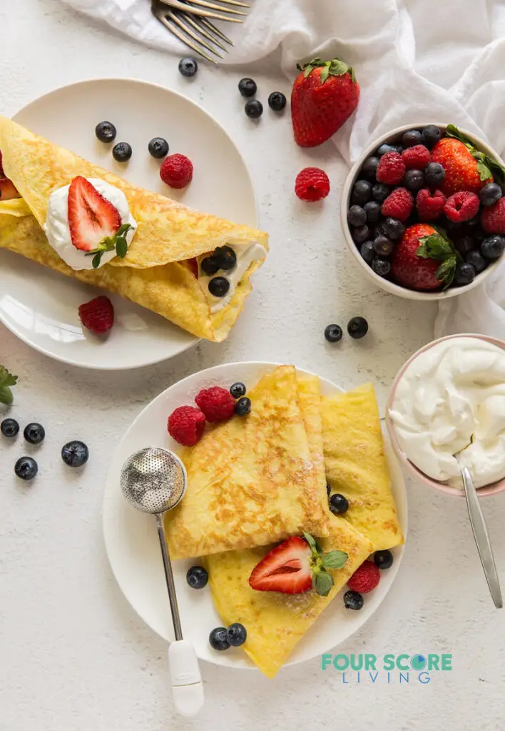 top down view of a countertop with one filled crepe on a plate, one plate of folded crepes, a bowl of berries, a bowl of whipped cream, and servingware