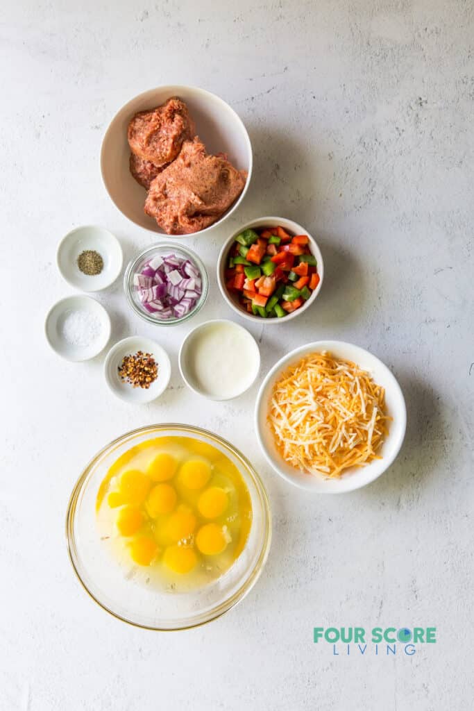 Top down view of ingredients to make breakfast casserole in separate bowls, including sausage, eggs, vegetables, cheese, and seasonings.