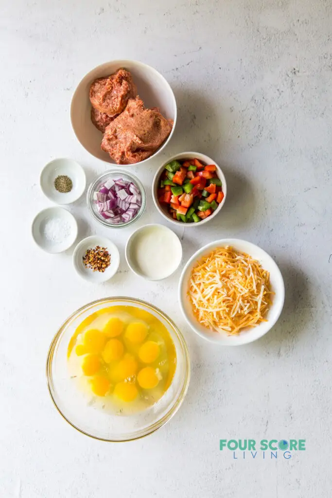 Top down view of ingredients to make breakfast casserole in separate bowls, including sausage, eggs, vegetables, cheese, and seasonings.