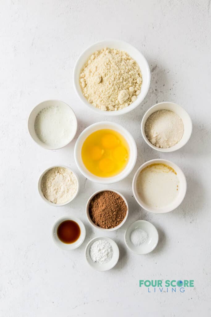 ingredients for keto chocolate cake all in separate small white bowls, including eggs, dry ingredients, sweeteners, vanilla, and cocoa.