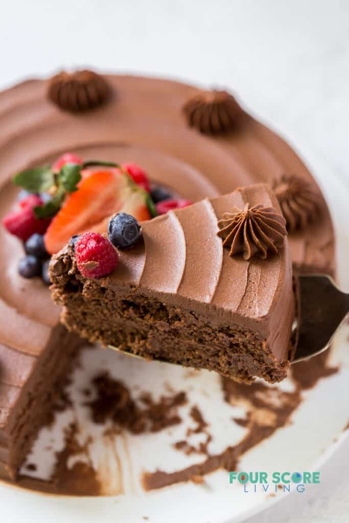 close up view of a slice of chocolate cake with a raspberry and blueberry on top. The cake slice is being lifted from a whole chocolate cake.