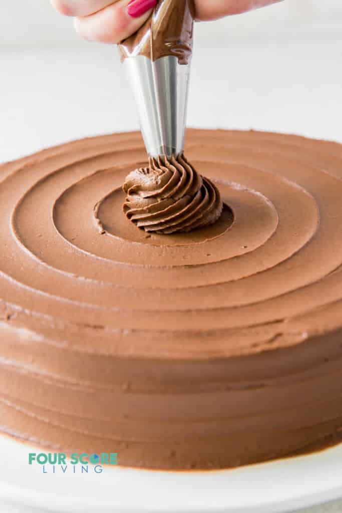 A chocolate cake being decorated with keto chocolate frosting in a piping bag in a swirl pattern.