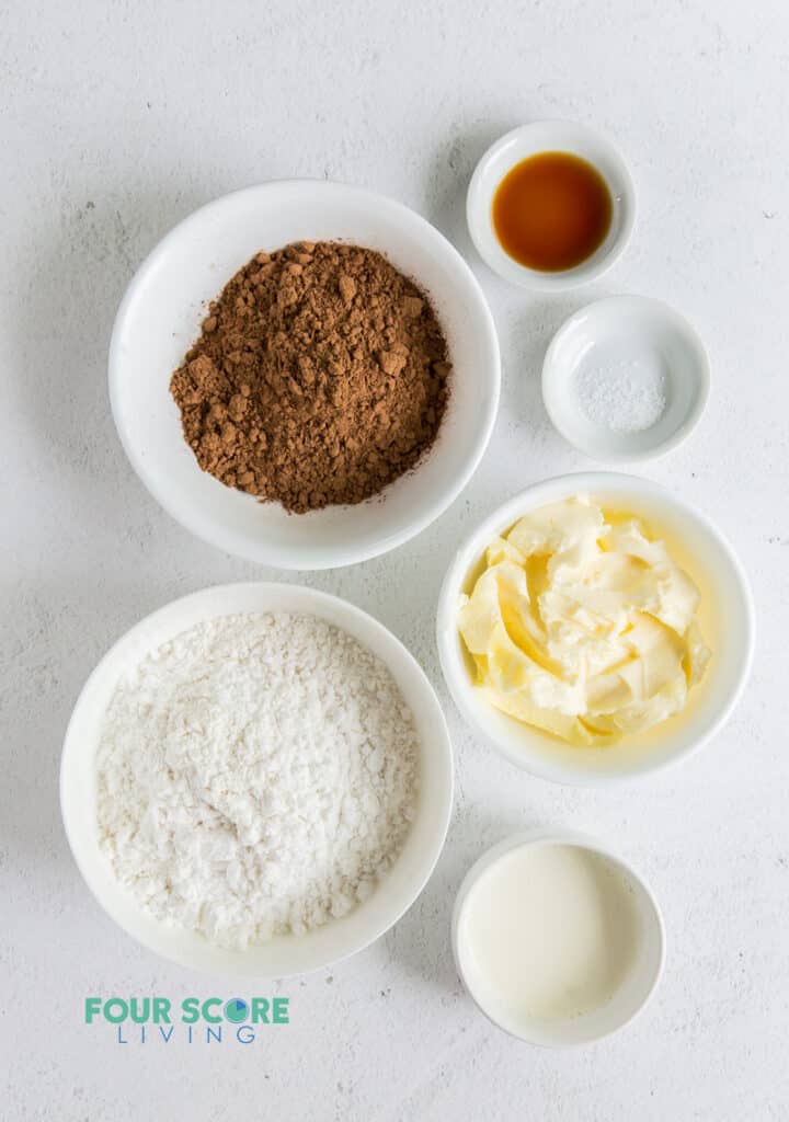 All of the ingredients for keto chocolate frosting in small white bowls. Ingredients include butter, cocoa, sweetener, cream, vanilla.