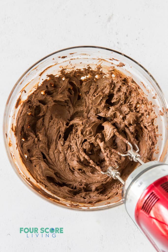 Chocolate frosting being mixed by a hand mixer in a glass bowl.
