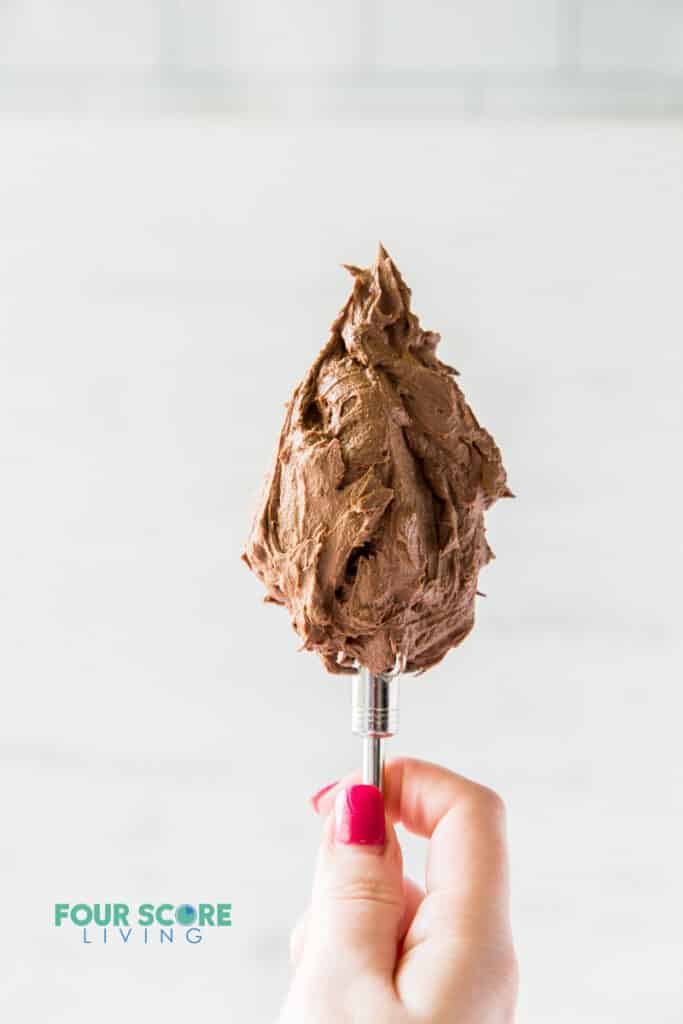 A mixer paddle full of keto chocolate frosting being held up in the air by a feminine hand with red nails.